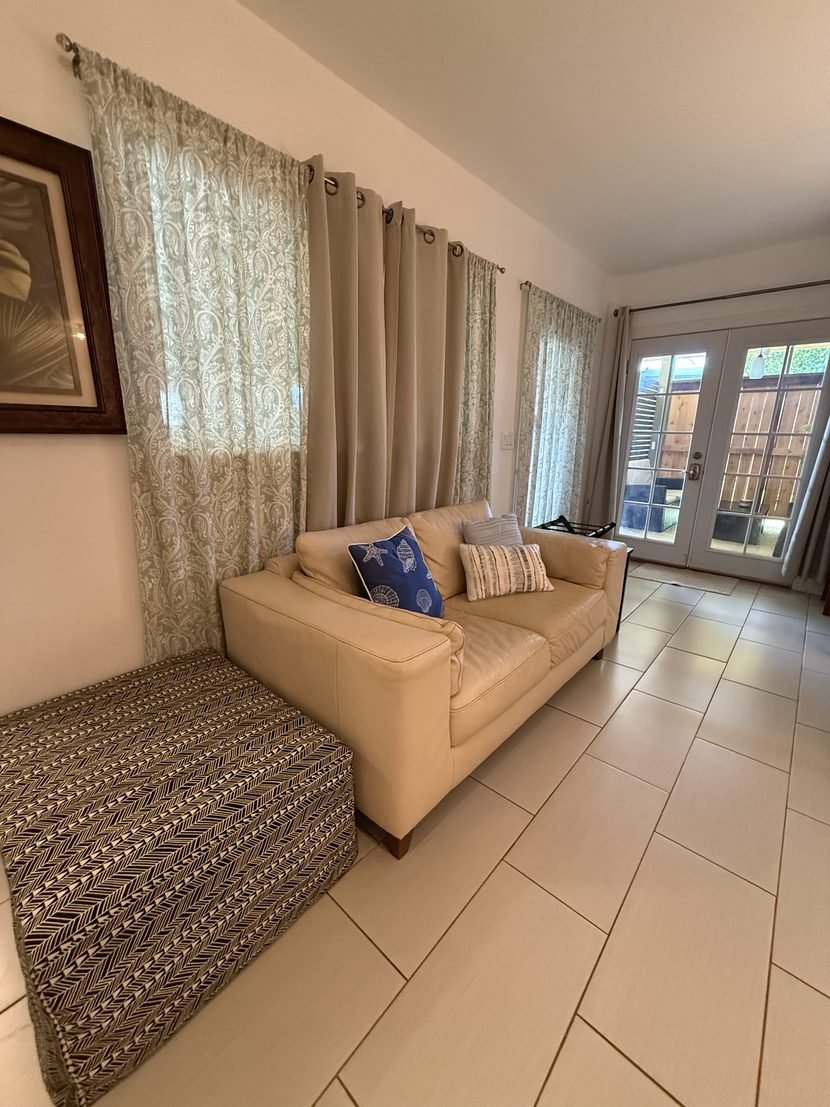 A tan sofa with cushions in the Sunrise studio, below sheer curtains and the French doors onto the lanai.