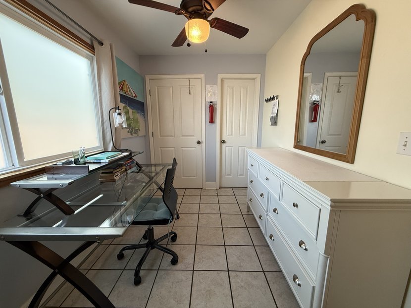 A glass desk and chair under a frosted window, beside a white dresser with an arched wood mirror, under a ceiling fan.